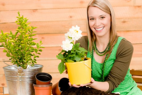 A smiling woman with blonde hair wearing a green polka-dot top and brown sleeves tends a small garden in an outdoor area with a wooden fence backdrop, showcasing a variety of potted plants. In front of her, she holds a bright yellow pot containing a healthy flowering plant with white blossoms and lush green foliage. To her left, there is a larger, silver metal pot with a green, bushy shrub, and in front of that, several empty flower pots in black and terracotta colors are arranged on a wooden surface. The garden space features a well-maintained grassy lawn with dense, vibrant green grass, and the overall scene appears to be set in a backyard or front garden area during daylight, suggesting a mild, pleasant weather condition. This natural yard setup highlights practices related to gardening and garden maintenance suitable for residential outdoor environments, reflecting the services offered by Garden Maintenance Alperton in the Alperton area, with a focus on healthy plant cultivation, container gardening, and outdoor beautification.