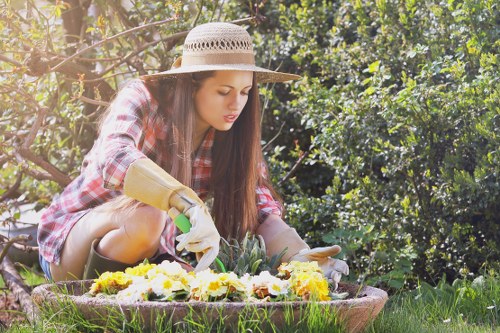 A young woman with long dark hair, wearing a wide-brimmed straw hat, a red and black checkered shirt, and gardening gloves, is kneeling in a lush garden. She is tending to a colorful flower bed that contains yellow, white, and purple blooms. The garden features a neatly maintained lawn with dense green shrubs and small trees in the background, under bright natural sunlight. The scene reflects careful outdoor maintenance typical of residential gardening services in Alperton, with the woman focused on planting or arranging flowers in an outdoor front or back garden space.